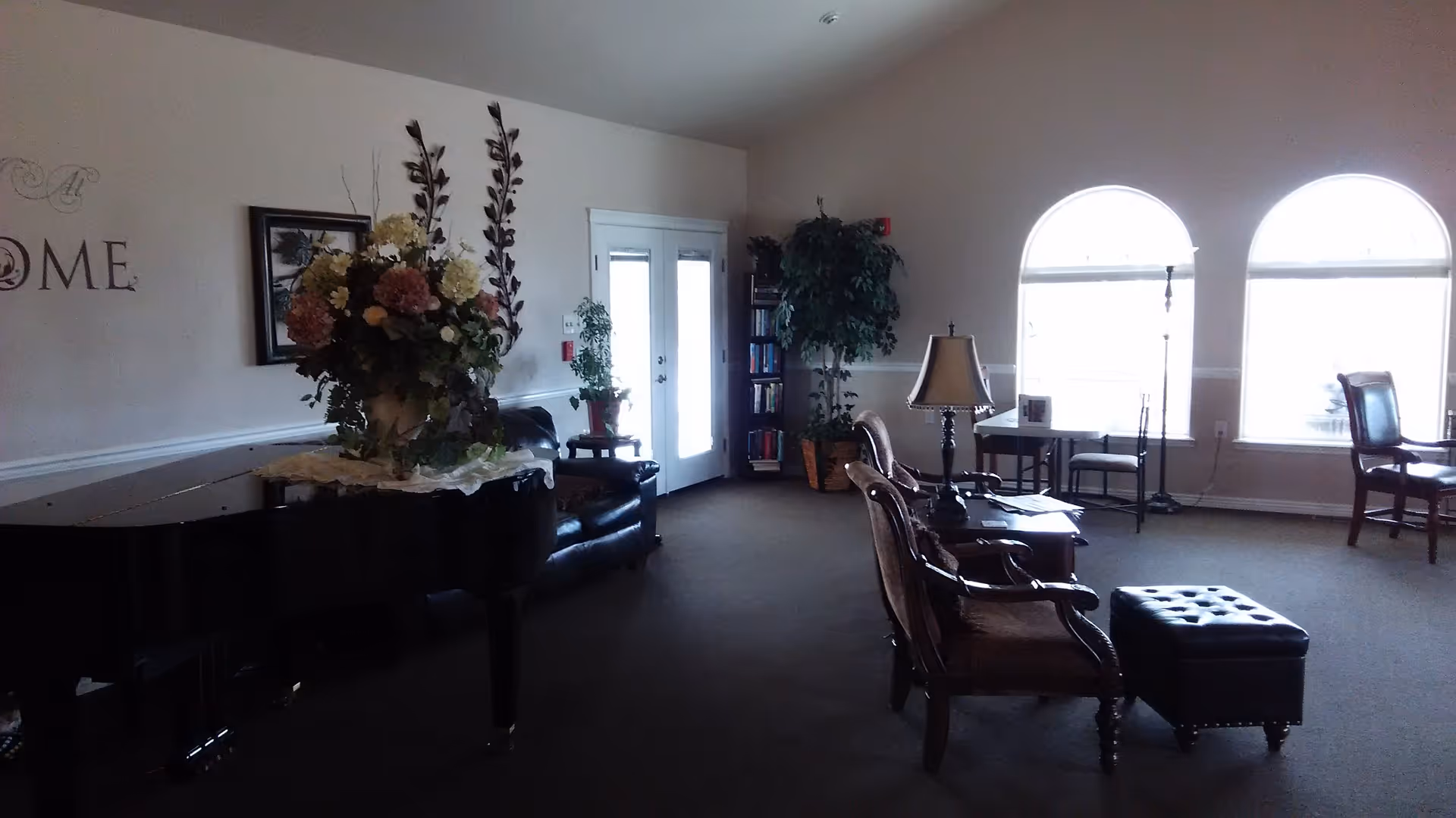 Spacious common room with a grand piano topped by a floral arrangement, cushioned chairs and ottoman, bookshelves, and arched windows.