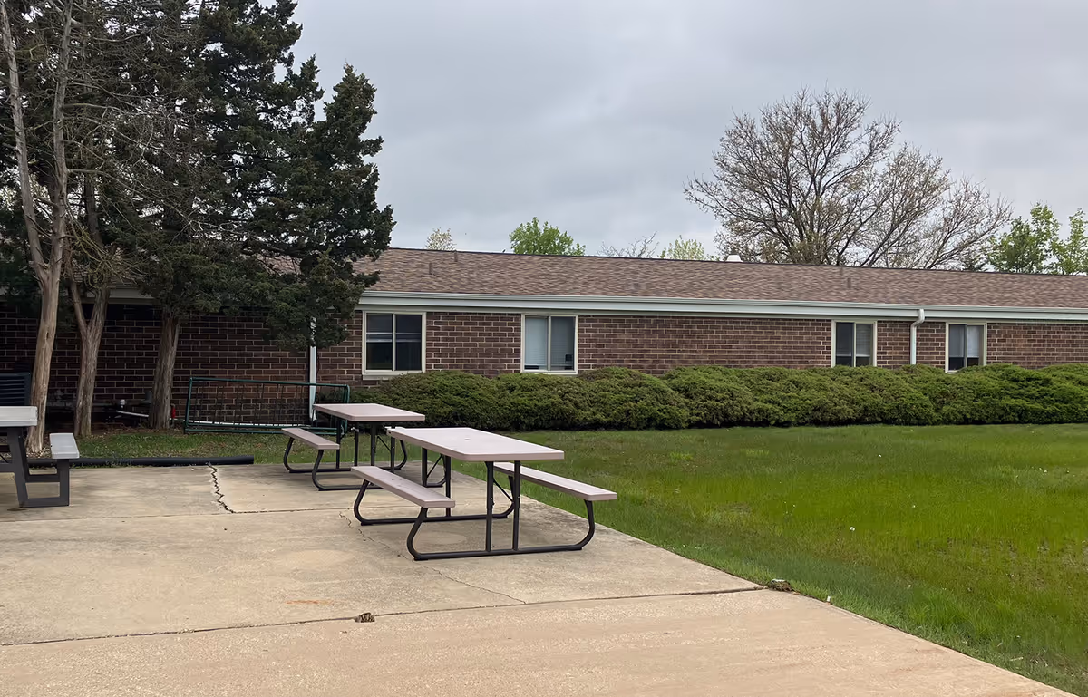 Outdoor patio area with two picnic tables and benches on a concrete surface next to a grassy lawn. In the background, there is a single-story brick building with several windows and some bushes along the base. Trees are visible behind the building under a cloudy sky.