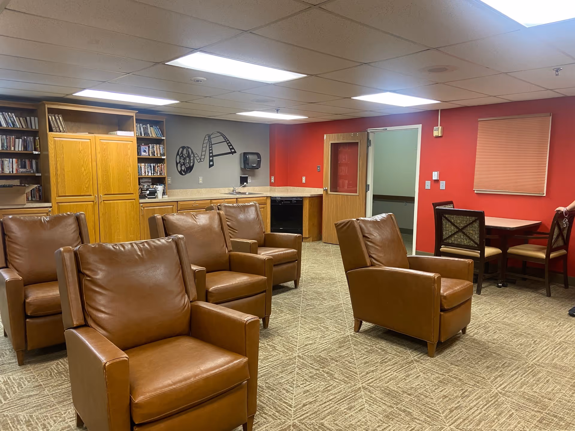 Communal lounge with several brown leather armchairs, bookshelves and cabinets, and a small table against a red wall.