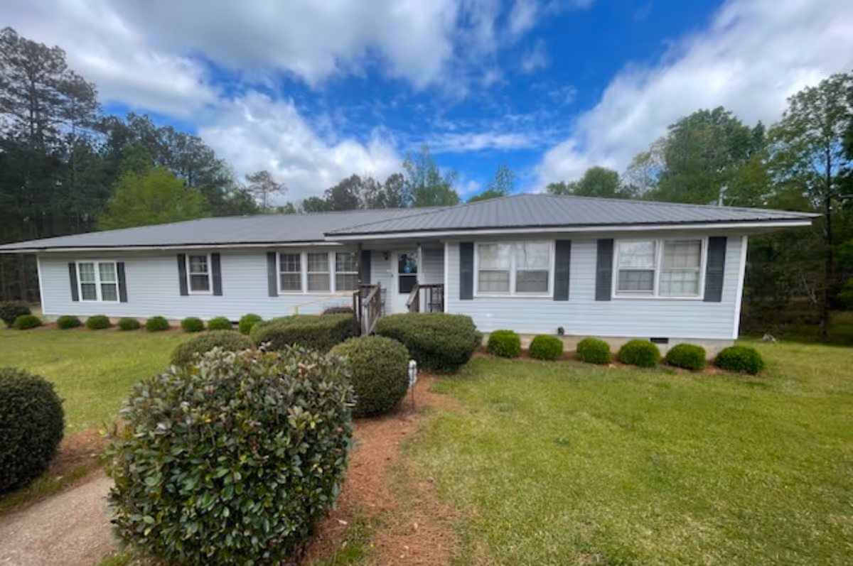 Single-story light blue house with a metal roof, front porch ramp, and manicured shrubs on a green lawn under a partly cloudy sky.
