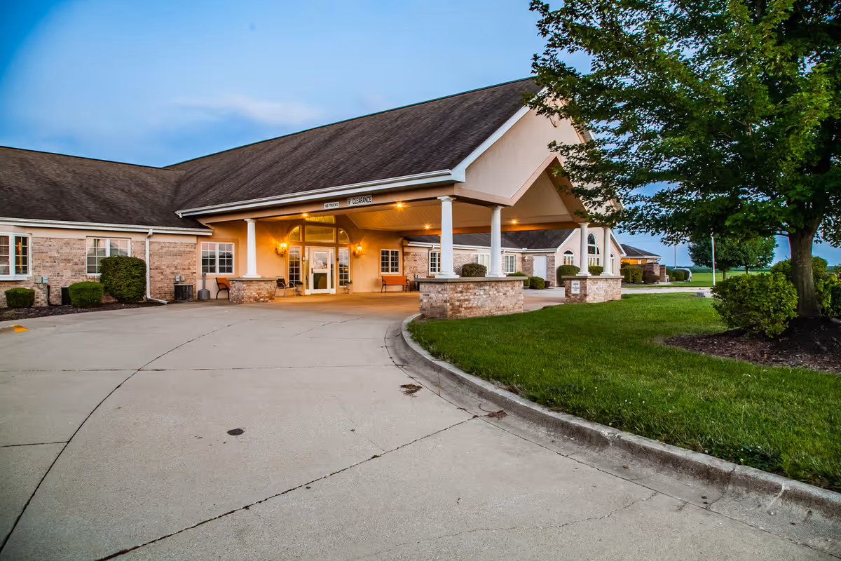 Exterior view of Brookstone Estates of Mattoon South showing the main entrance with a covered driveway, brick and beige walls, outdoor lighting, and surrounding greenery including trees and bushes.