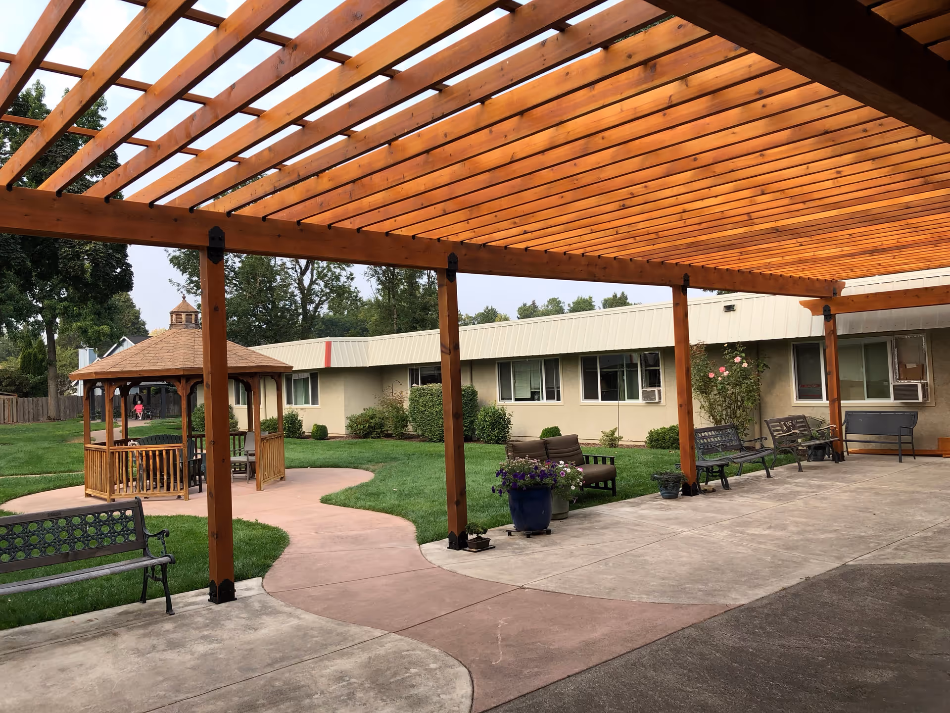Wooden pergola-covered courtyard with benches, a gazebo, potted plants, and a single-story senior living building in the background.