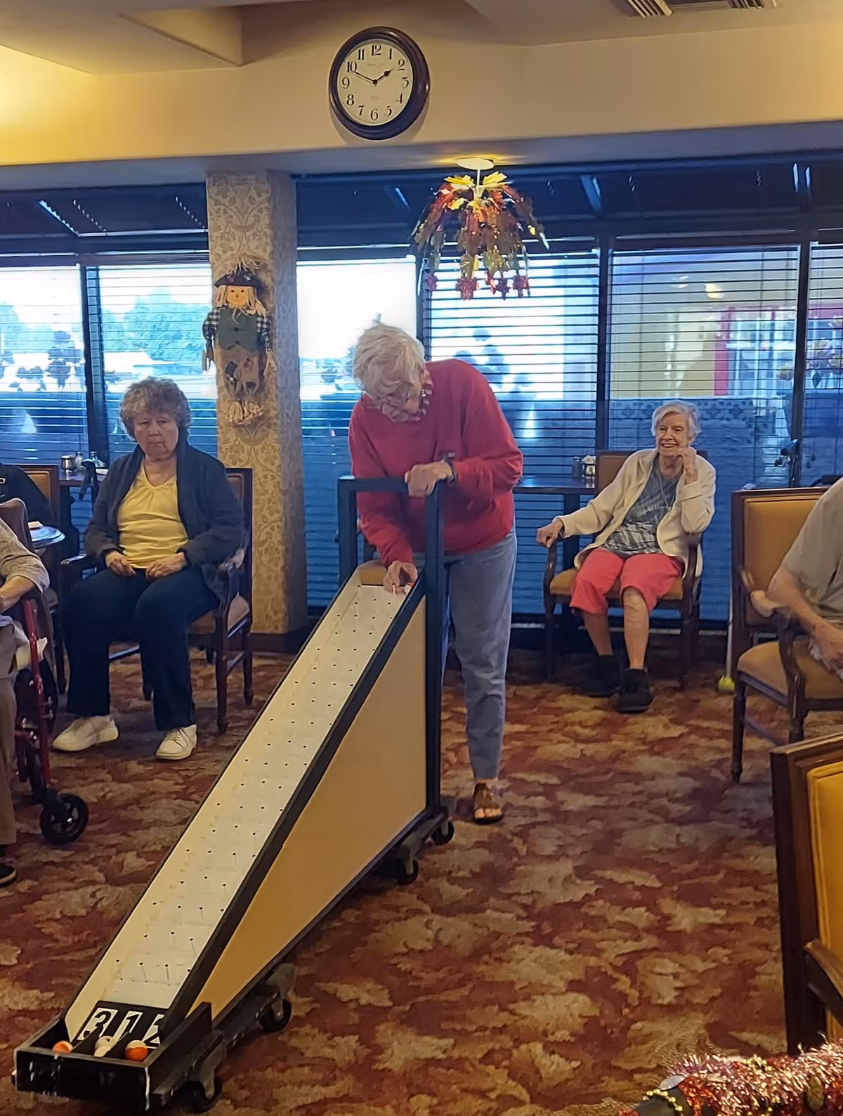 A group of elderly people sitting and standing in a common room with carpeted floor and large windows. One elderly woman in a red sweater is playing a tabletop bowling game while others watch and sit in chairs. A clock on the wall shows the time as 11:10.