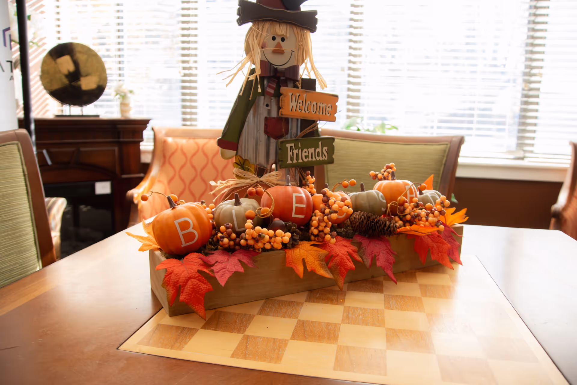 Autumn-themed centerpiece with mini pumpkins, berries and a 'Welcome Friends' scarecrow on a dining table in a sunlit room.