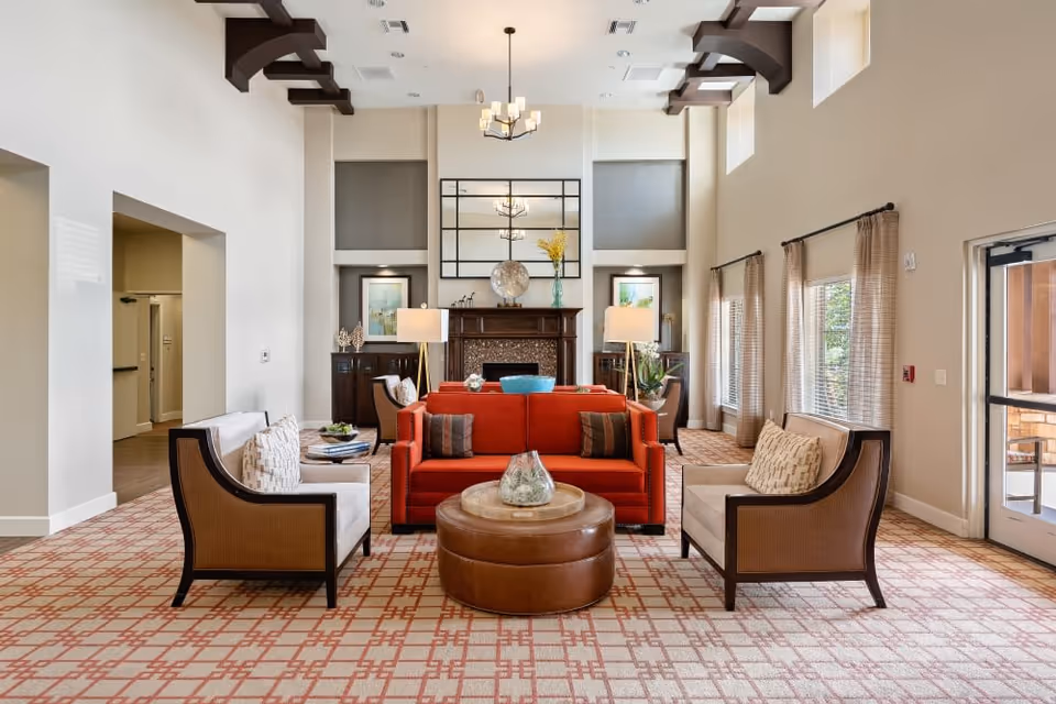 A spacious and well-lit living room area with a red sofa, two beige armchairs, a round brown ottoman, and a fireplace with a large mirror above it. The room features high ceilings with exposed wooden beams, large windows with curtains, and decorative lamps on either side of the fireplace.
