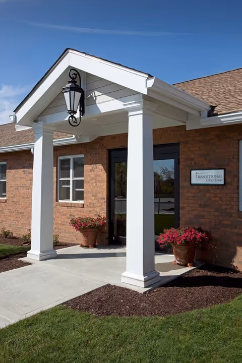 Front entrance of a brick building with white columns, a covered portico, a hanging lantern, and potted flowers.