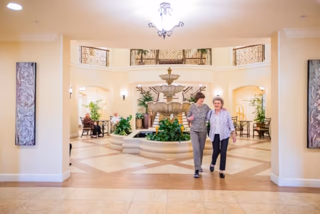 Two elderly women walking arm in arm through a spacious, well-lit lobby area with a large decorative fountain in the center, surrounded by plants and seating areas. The lobby features beige walls, tiled floors, and ornate railings on the upper level. Two other elderly individuals are seated in the background near the walls.