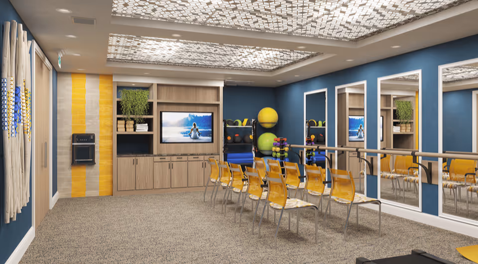 Bright interior activity/exercise room with rows of yellow chairs facing a TV, mirrored wall, ballet barre and exercise equipment.