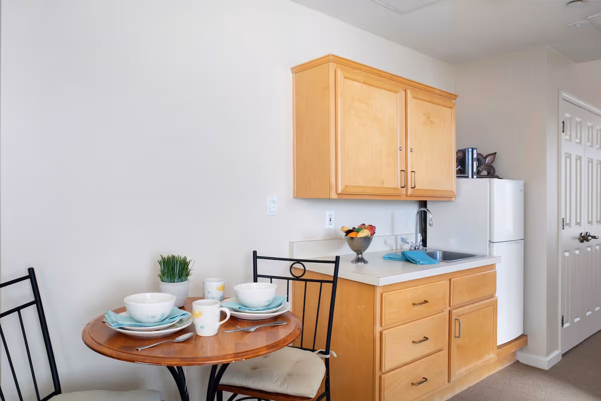 Small kitchen area with light wood cabinets, a white countertop, a sink, and a white refrigerator. Next to the kitchen is a round wooden table set for two with bowls, plates, mugs, and silverware. There is a small potted plant on the table. The walls are light-colored and there is a door with white double panels in the background.