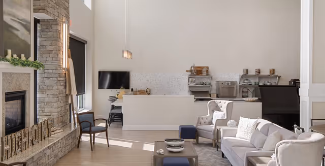 Bright communal living room with stone fireplace, sofas and armchairs around a coffee table and a small kitchenette counter in the back.