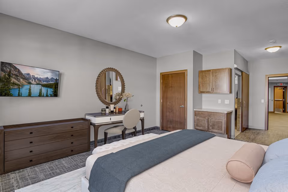 A senior living facility bedroom featuring a bed with pillows and a blanket, a wooden dresser with a mounted flat-screen TV above it, a vanity table with a round mirror and a chair, and wooden cabinets on the wall. The room has neutral-colored walls and carpeted flooring, with a door leading to a hallway.