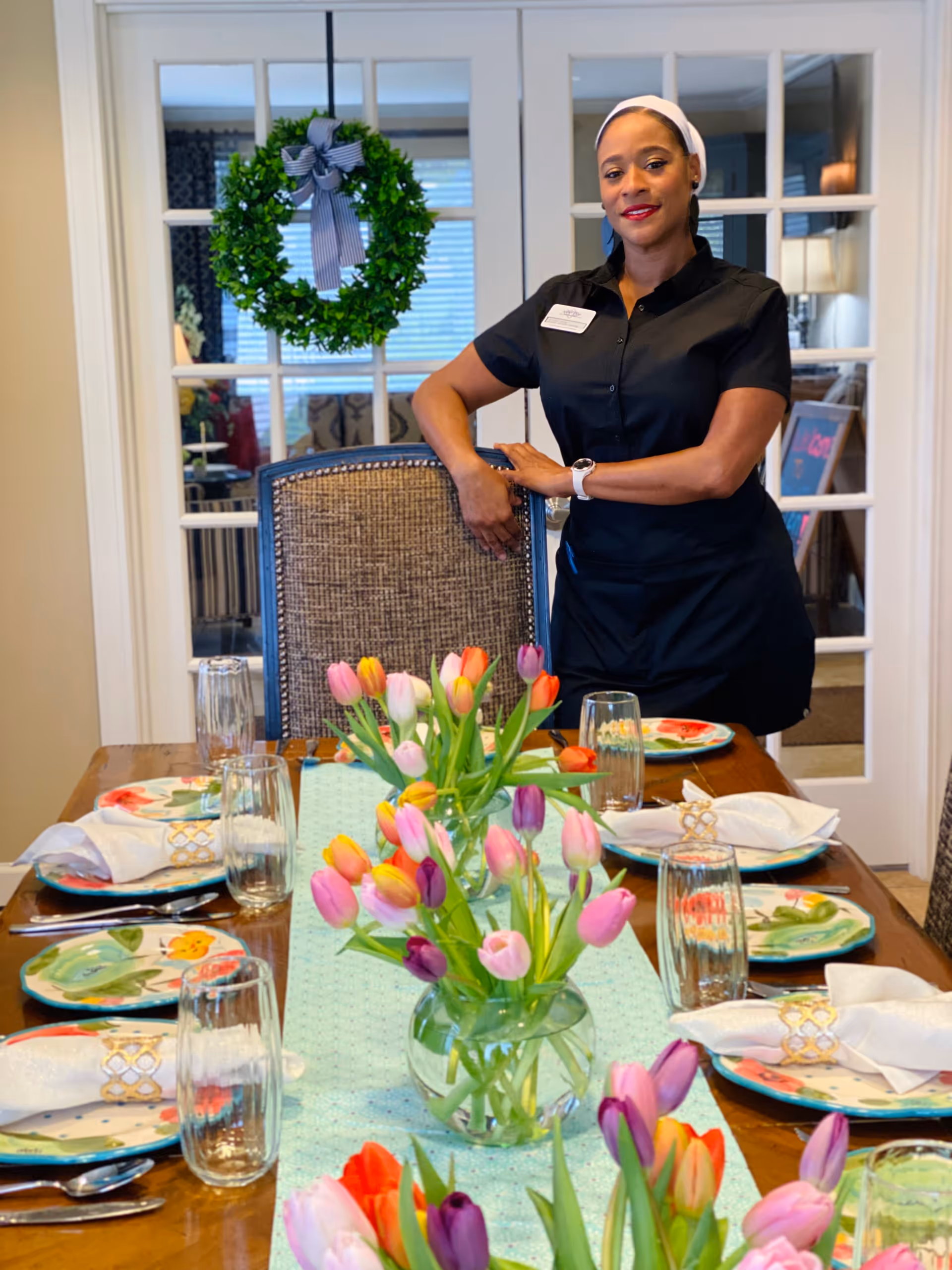 A woman dressed in a black uniform stands behind a dining chair at a dining table set with colorful plates, napkins, and glasses. The table is decorated with vases of vibrant tulips. Behind her are glass-paneled double doors with a green wreath hanging on one of them.