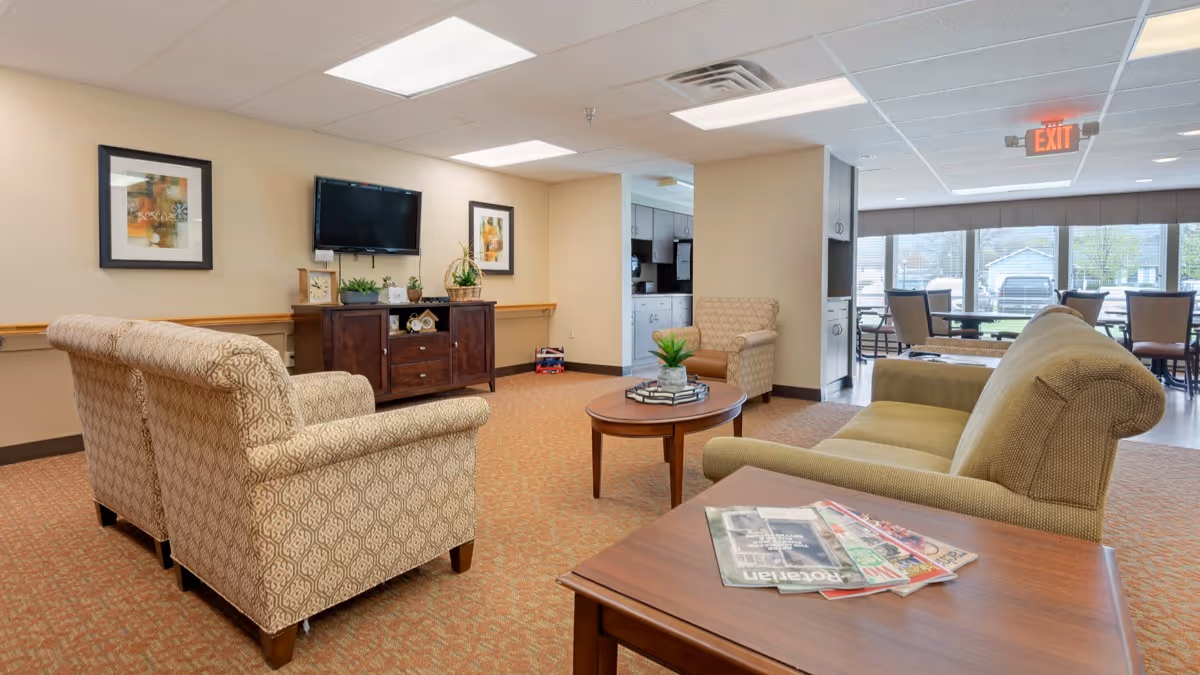 A bright senior living lounge with upholstered chairs, a sofa, coffee tables, a wall-mounted TV, and a dining area visible through large windows.