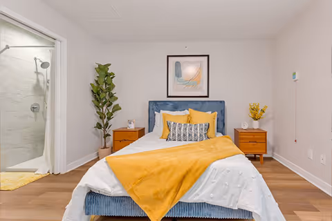 A neatly made bed with a blue upholstered headboard, white bedding, and a yellow throw blanket in a bedroom. There are two wooden nightstands on either side of the bed, one with a small plant and the other with a clock and a vase with yellow flowers. A framed abstract artwork hangs on the wall above the bed. To the left, there is an open doorway leading to a bathroom with a glass shower enclosure.