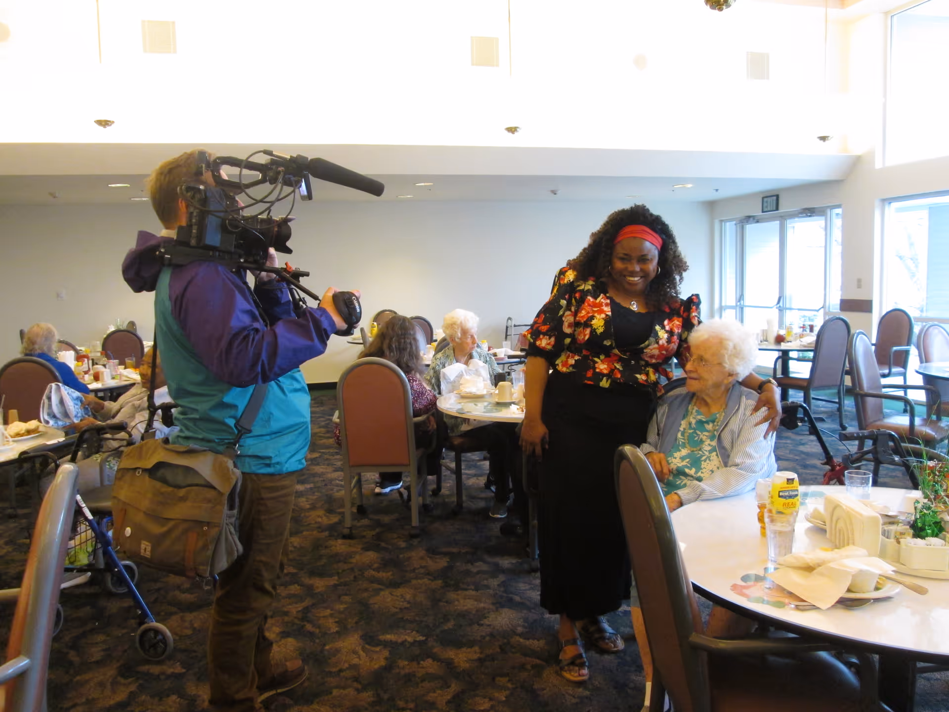 A woman with a floral top and red headband smiles and stands next to an elderly woman seated at a dining table in a senior living facility. A cameraman is filming them, and other elderly residents are seated at tables in the background.