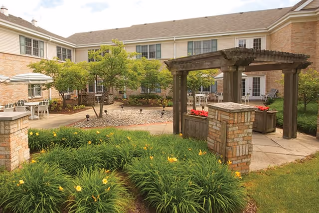 Outdoor courtyard area of a senior living facility with green shrubs and yellow flowers in the foreground, a stone and wood pergola structure, paved walkways, small trees, and a two-story building with beige siding and brick exterior in the background.