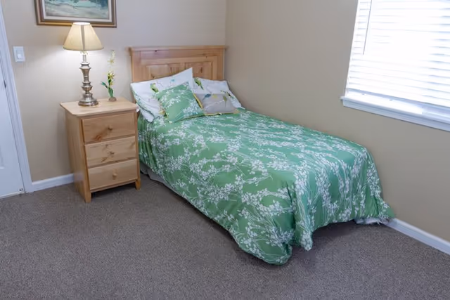 A small bedroom with a single bed covered in a green floral bedspread. There are several pillows on the bed, and a wooden nightstand with three drawers beside it. On the nightstand is a lamp and a small flower arrangement. A window with white blinds is on the right wall, and a framed picture hangs above the nightstand.