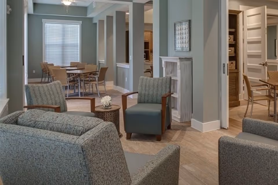 A cozy interior common area with several upholstered armchairs arranged around a small round table with a flower decoration. In the background, there is a dining area with multiple tables and chairs near a window with blinds. The walls are painted a soft grayish-blue, and there is a white decorative fireplace and a framed artwork on the wall.