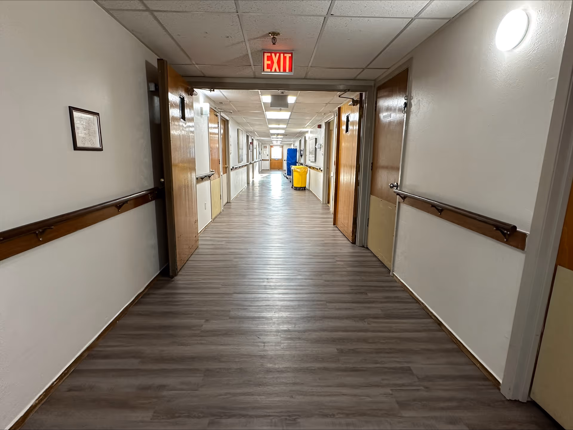 A long, empty hallway in a nursing and rehab facility with wooden doors on both sides, handrails along the walls, a lighted exit sign at the end, and a yellow cleaning cart visible in the distance.