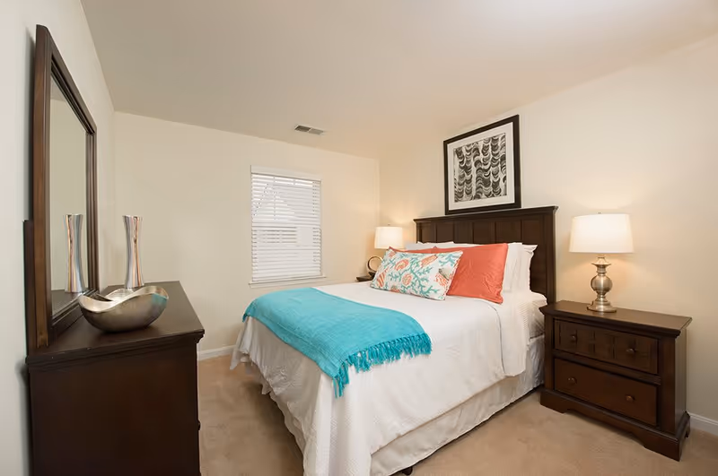 Well-lit bedroom with a bed dressed in white linens, a teal throw and decorative pillows, dark wood headboard, matching nightstands and dresser with a mirror.