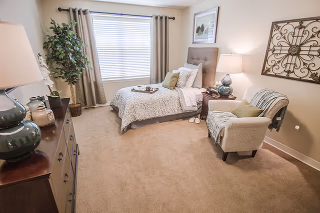 A cozy bedroom with a single bed dressed in patterned bedding and multiple pillows. Next to the bed is a nightstand with a lamp and decorative items. A comfortable armchair with a throw blanket and pillow is placed nearby. The room has beige carpeting, a large window with curtains, a dresser with a lamp and decor, a potted plant, and framed artwork on the walls.