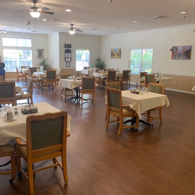 A spacious dining room with multiple tables covered with white tablecloths, each set with glasses and condiments. Wooden chairs with cushioned backs surround the tables. The room has large windows allowing natural light to fill the space, ceiling fans, and framed artwork on the walls.