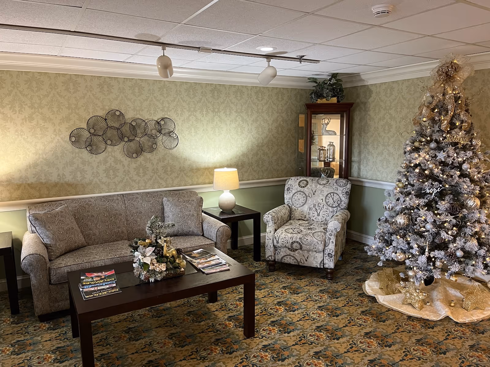 A cozy living room with a patterned sofa and armchair, a wooden coffee table with magazines and a decorative centerpiece, a side table with a lamp, a glass display cabinet with decorative items, and a decorated Christmas tree with gold and silver ornaments and lights.