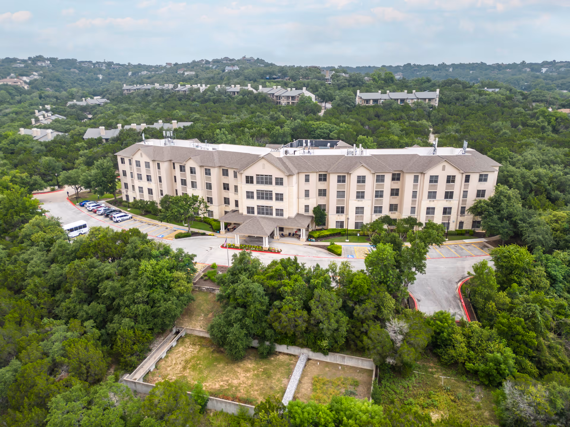 Aerial view of a large, multi-story senior living facility surrounded by dense green trees and hills. The building has a beige exterior with multiple windows and a covered entrance. Several cars are parked in the parking lot adjacent to the building.