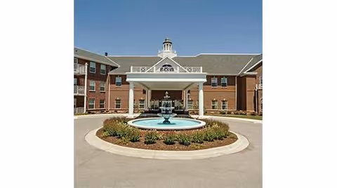 Front exterior view of a large brick senior living facility with a covered entrance and a circular driveway featuring a central water fountain surrounded by landscaping.