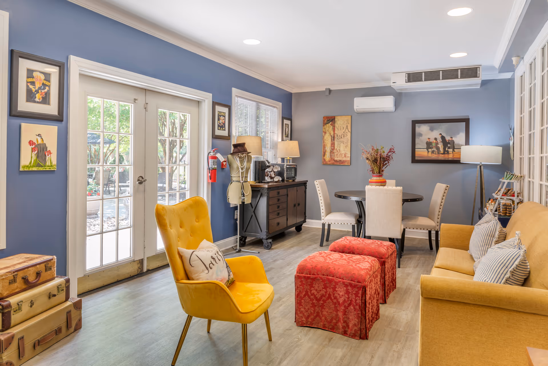A cozy living room area with blue walls, a yellow armchair with a Paris-themed pillow, two red ottomans, a mustard yellow sofa with striped pillows, a round dining table with four beige chairs, and various wall art. French doors lead outside to a garden area, and there is a sideboard with a lamp, a coffee maker, and a mannequin adorned with necklaces and a hat.