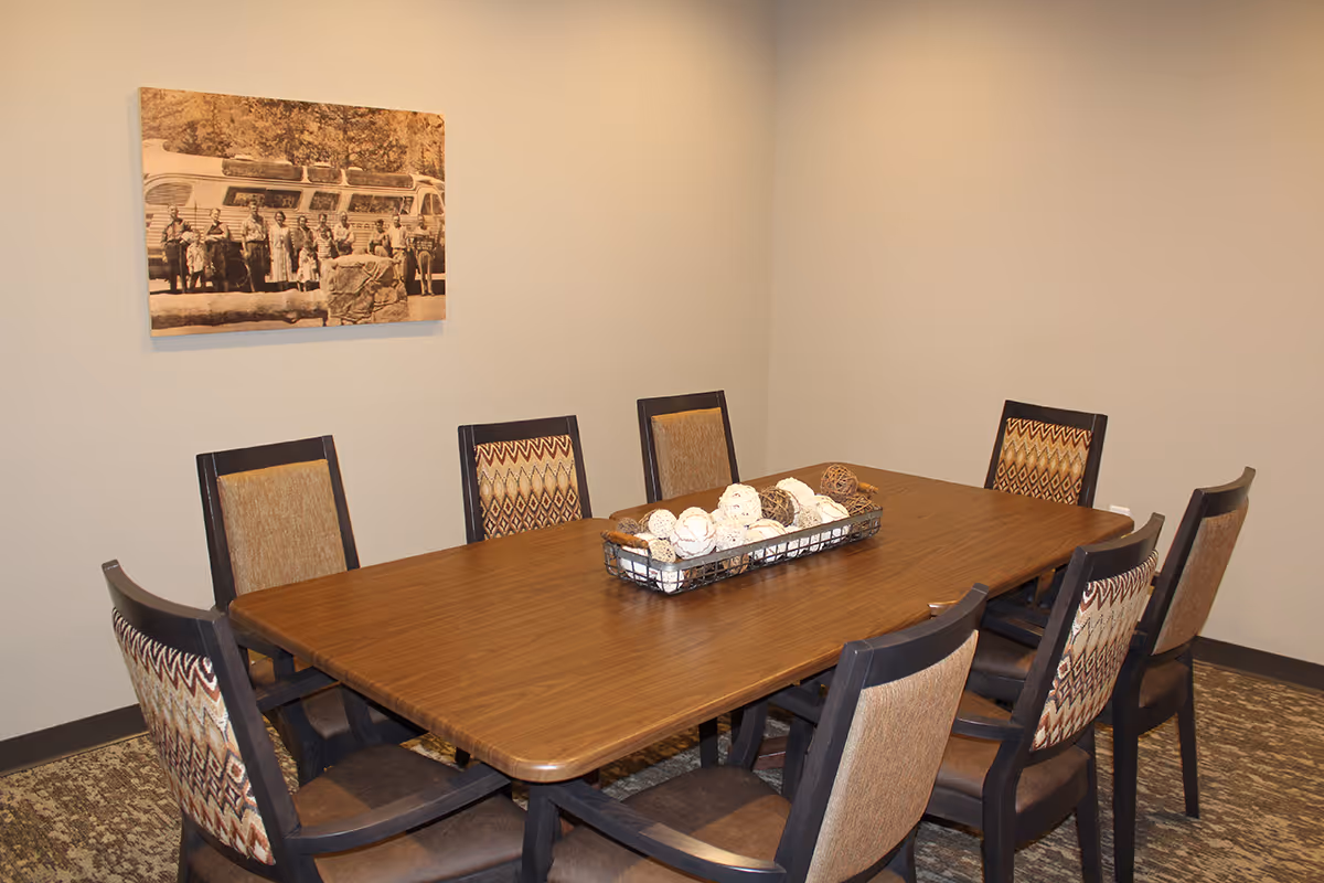 A dining room with a rectangular wooden table surrounded by eight chairs with patterned upholstery. A decorative centerpiece with assorted spherical objects is placed on the table. A sepia-toned photograph hangs on the beige wall behind the table.