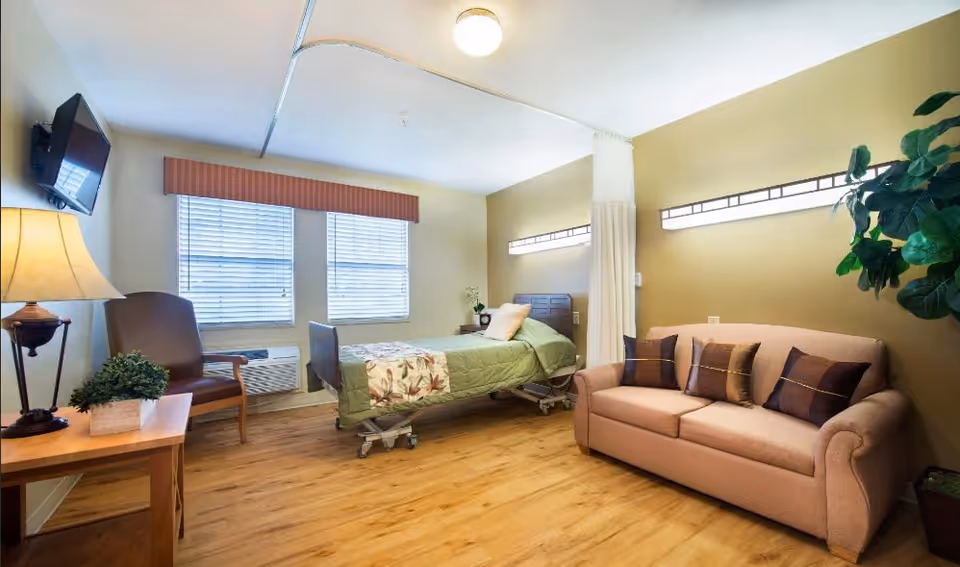 A well-lit room in a healthcare facility featuring a hospital bed with green bedding, a beige sofa with decorative pillows, a wooden chair, a side table with a lamp and plant, two windows with blinds, and a mounted flat-screen TV. The room has wooden flooring and light-colored walls with modern lighting fixtures.