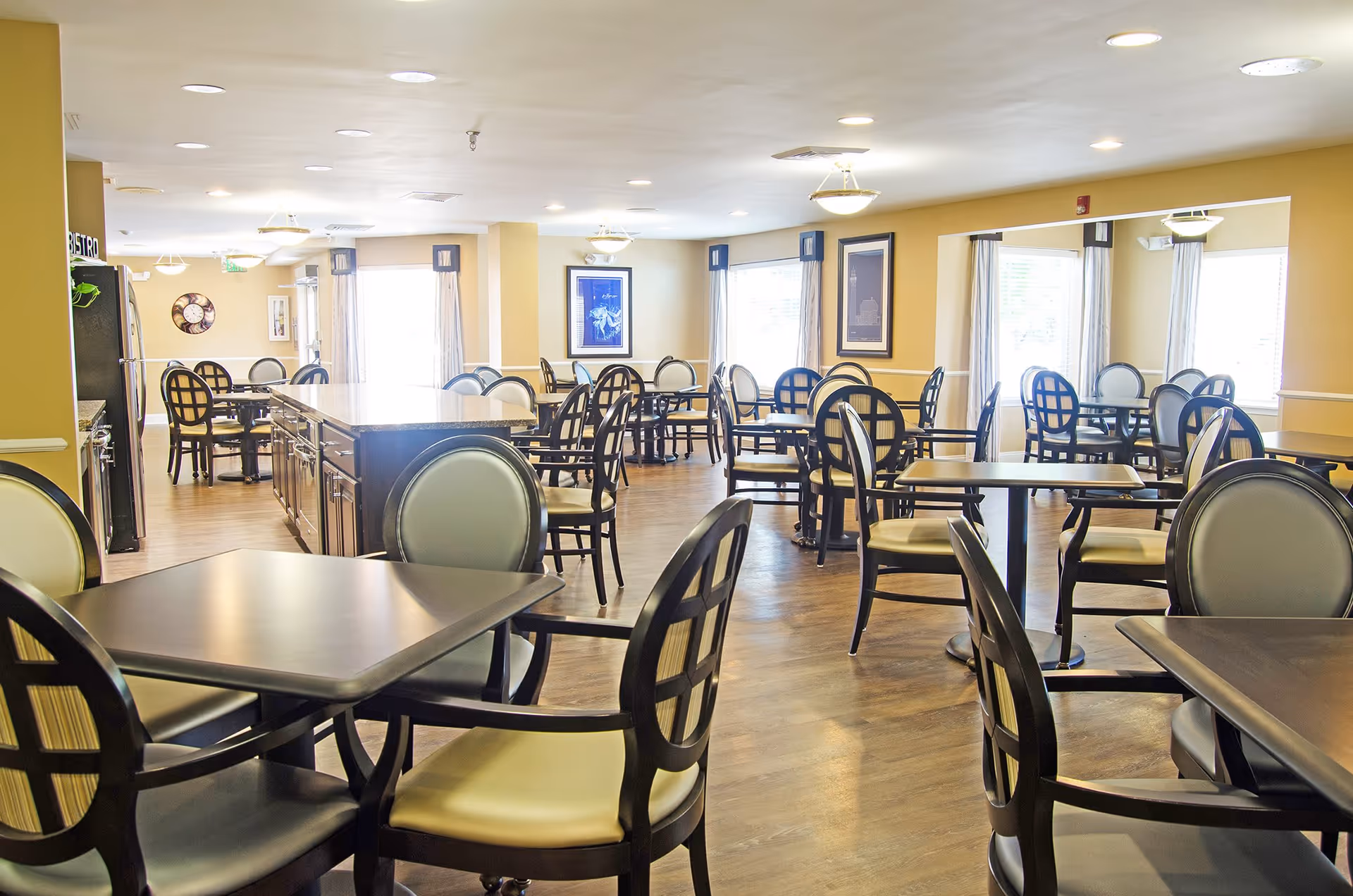 Bright and spacious dining room with multiple tables and chairs arranged neatly. The room has large windows with curtains allowing natural light to fill the space. The walls are painted a warm yellow color and decorated with framed artwork. The floor is wooden, and there is a large central island or counter in the middle of the room.