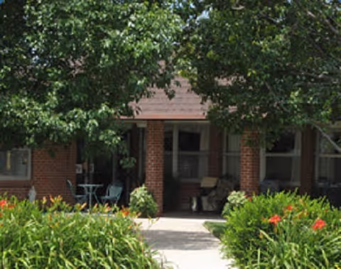 View of the front entrance of a brick building partially obscured by large green trees and bushes with orange flowers. There is a small patio area with a table and chairs near the entrance.