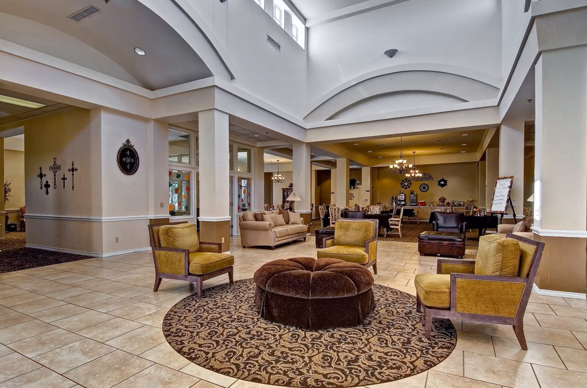A spacious and well-lit common area in a senior living facility featuring a circular patterned rug with a large round brown ottoman in the center, surrounded by four mustard-yellow cushioned armchairs. In the background, there are beige sofas, dining tables with chairs, chandeliers, and decorative wall art including crosses and clocks. The room has high ceilings with architectural arches and large windows near the ceiling allowing natural light.