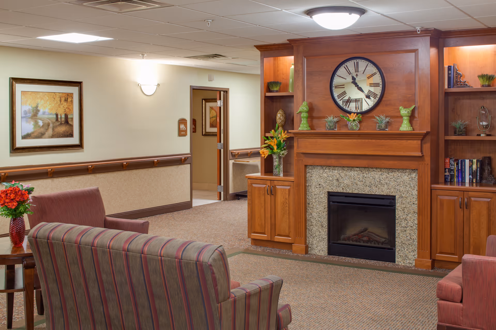 Cozy common room with a wood-paneled fireplace and clock, built-in shelves, and striped armchairs.