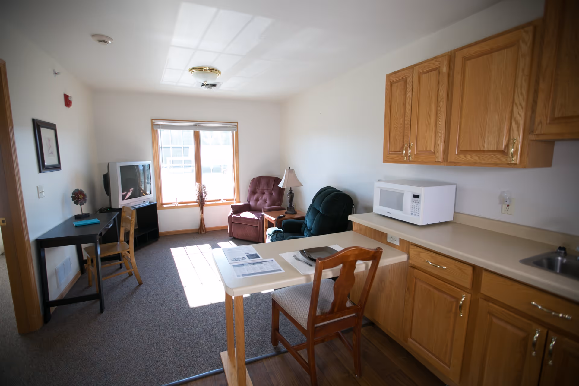 A small senior living facility room with a kitchenette featuring wooden cabinets, a microwave, and a sink. There is a small dining counter with a chair and place setting. The room also includes a living area with two armchairs, a side table with a lamp, a desk with a chair, and a television near a window letting in natural light.