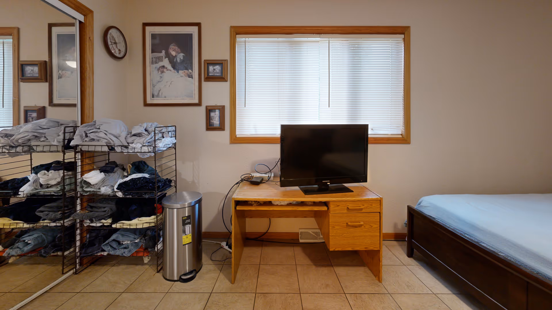 A bedroom with a wooden bed frame and light blue bedding on the right side. In the center, there is a wooden desk with a flat-screen TV on top. Behind the desk is a window with closed blinds. On the left side, there is a metal rack filled with folded clothes and a silver trash can next to it. The wall has a clock and framed pictures hanging above the desk and rack. A large mirror with a wooden frame is visible on the far left side.