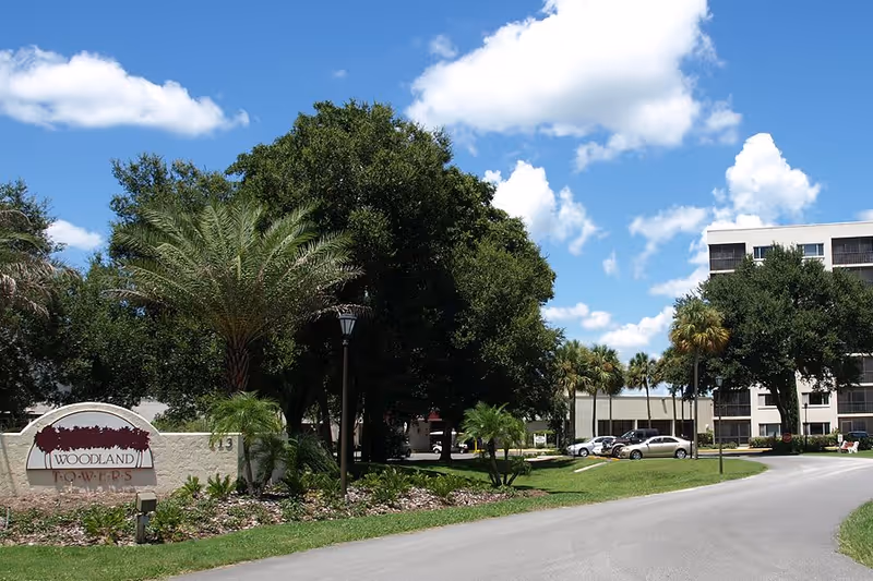 Exterior view of Woodland Towers senior living facility with a driveway, landscaped greenery including palm trees and bushes, and a multi-story building in the background under a blue sky with scattered clouds.
