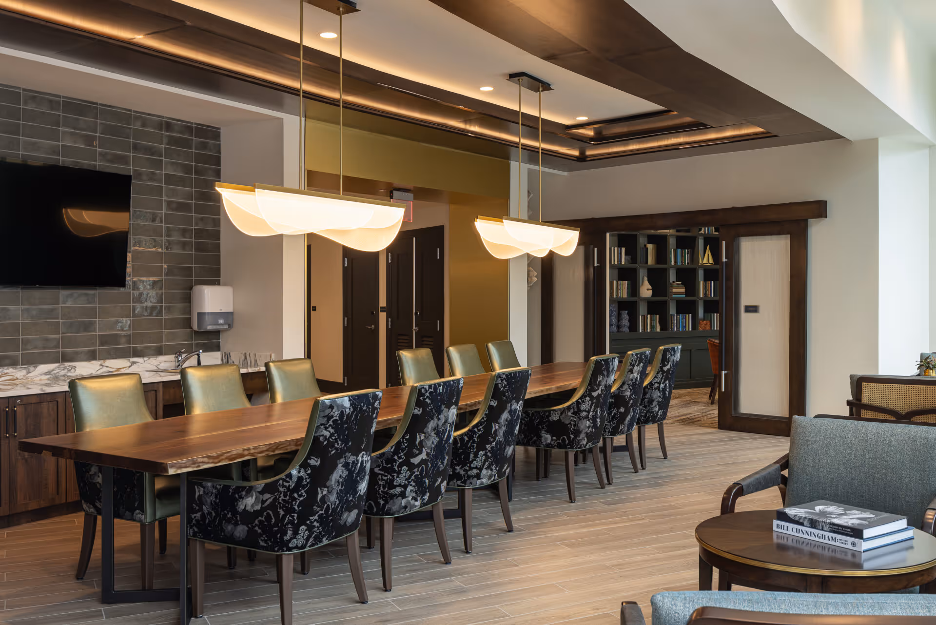 A modern dining room with a long wooden table surrounded by green and patterned chairs. Two large pendant lights hang above the table. In the background, there is a wall-mounted TV, a countertop with a marble surface, and a bookshelf filled with books. The room features wooden flooring and a mix of wood and white walls.