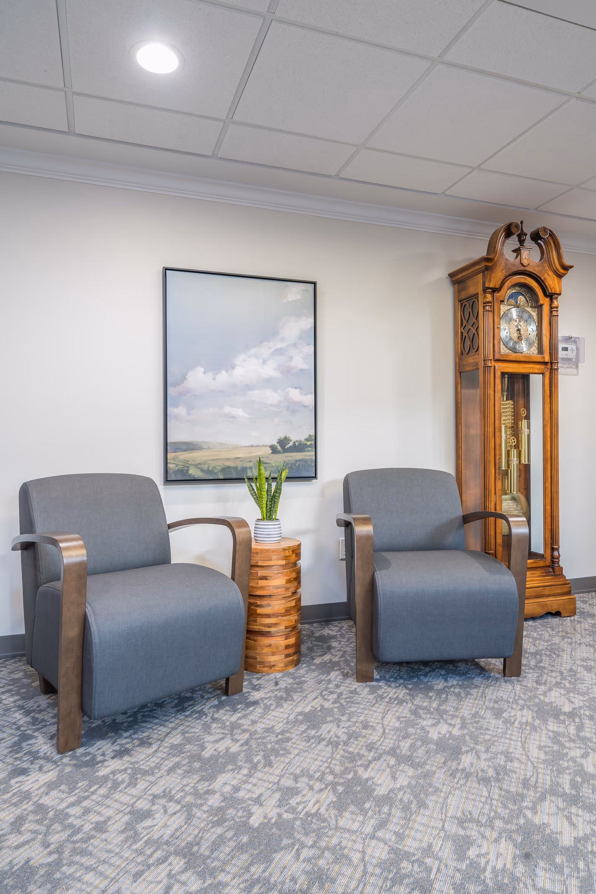 A seating area with two gray armchairs featuring wooden armrests, a small round wooden side table with a potted plant, a large framed landscape painting on the wall, and a tall wooden grandfather clock in the corner.