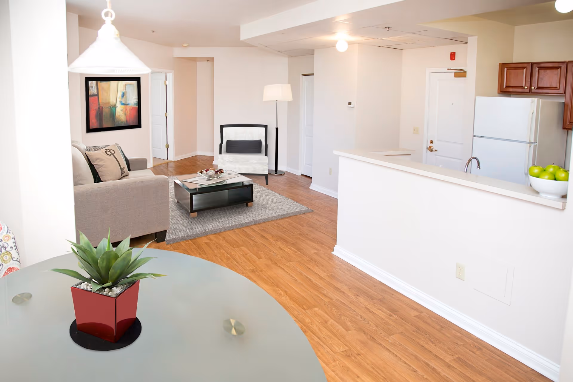 Interior view of a senior apartment living area with light wood flooring, a beige sofa with decorative pillows, a glass coffee table on a gray rug, a white armchair with a black cushion, a floor lamp, and a kitchen area with white appliances and wooden cabinets. A round glass table with a small green plant in a red pot is in the foreground.