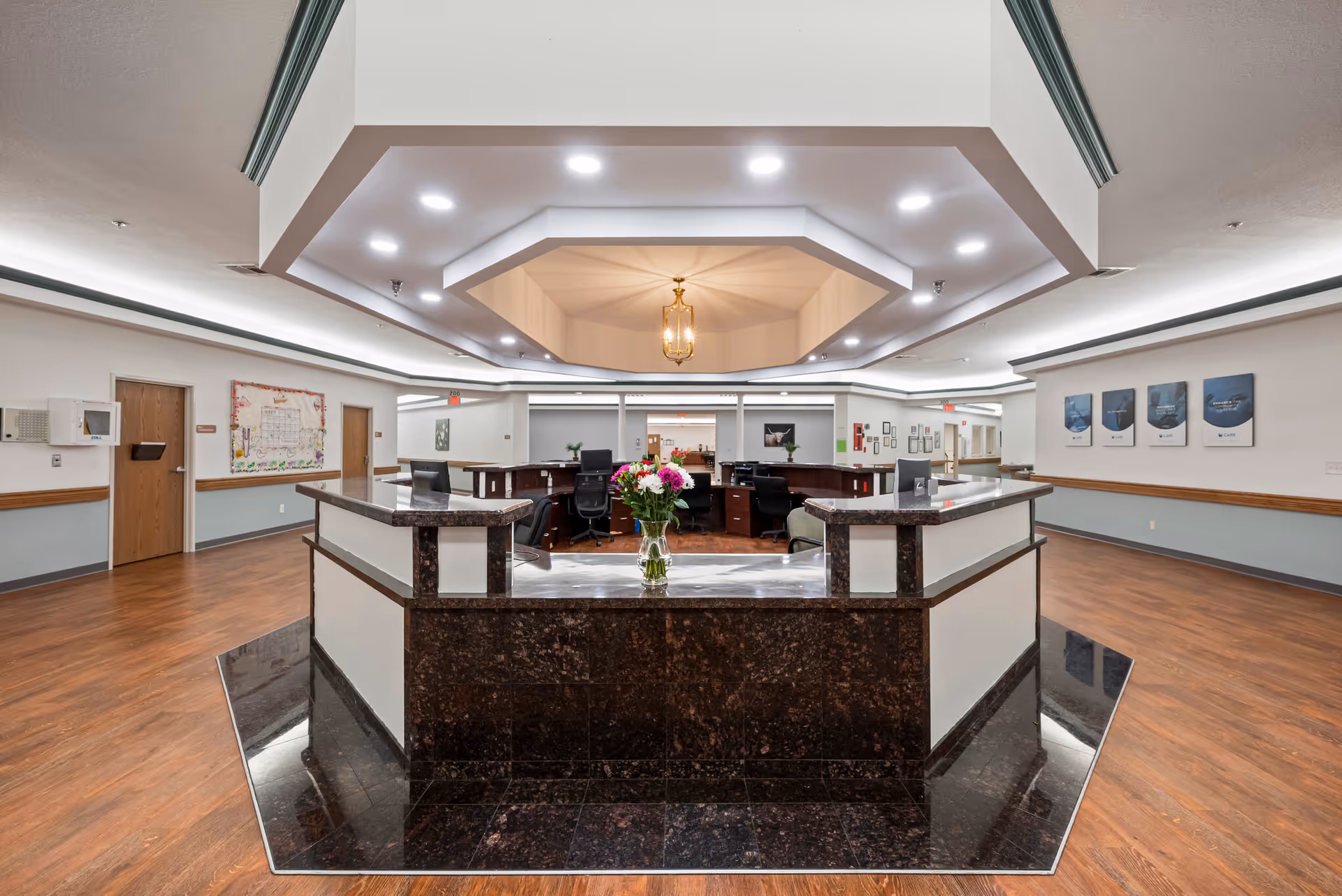 A spacious nursing facility reception area with a large, dark marble front desk in the center. The desk is surrounded by wood flooring and has a vase of colorful flowers on top. The ceiling features recessed lighting and a decorative octagonal light fixture. Several office chairs and desks are visible behind the reception desk, with doors and framed pictures on the walls in the background.