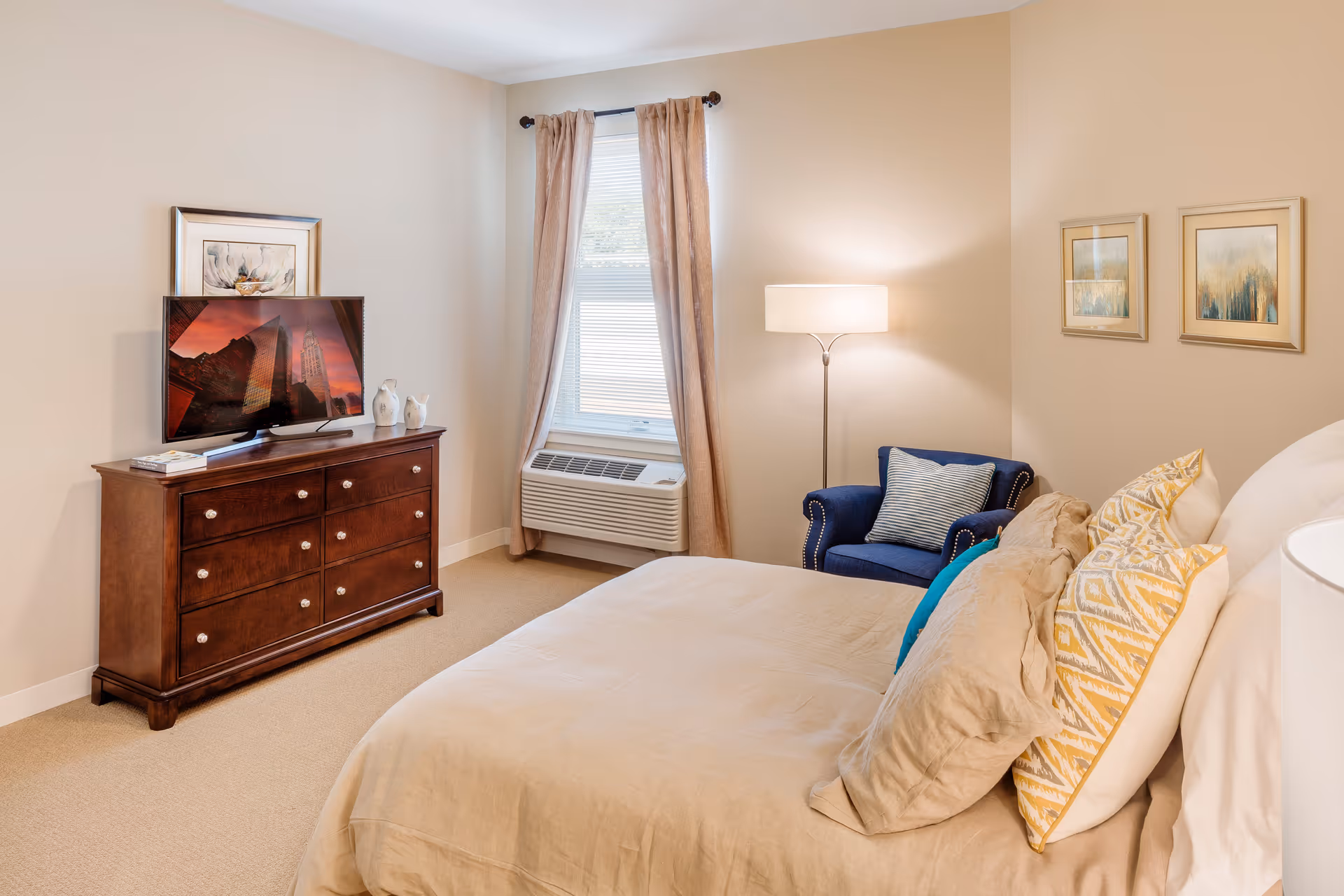 A cozy bedroom with a beige bedspread and multiple pillows, a dark wooden dresser with a flat-screen TV on top, a window with beige curtains, a blue armchair with a striped pillow, a floor lamp, and two framed pictures on the wall.