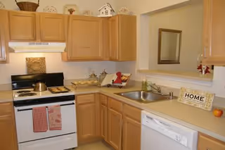A compact kitchen with light wood cabinets, a white stove with a kettle and baking tray, a countertop with decorative items including a small sign that says HOME, a sink, and a white dishwasher. There is a pass-through window with a mirror on the wall behind the sink.
