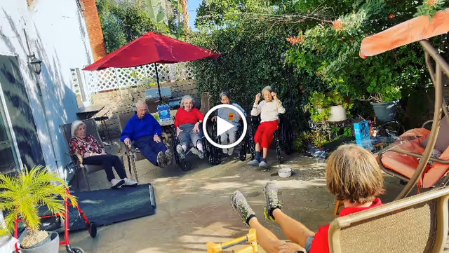 A group of elderly people sitting outdoors in a garden patio area, some in wheelchairs and others in chairs, enjoying the sunny weather with greenery and a red umbrella providing shade.