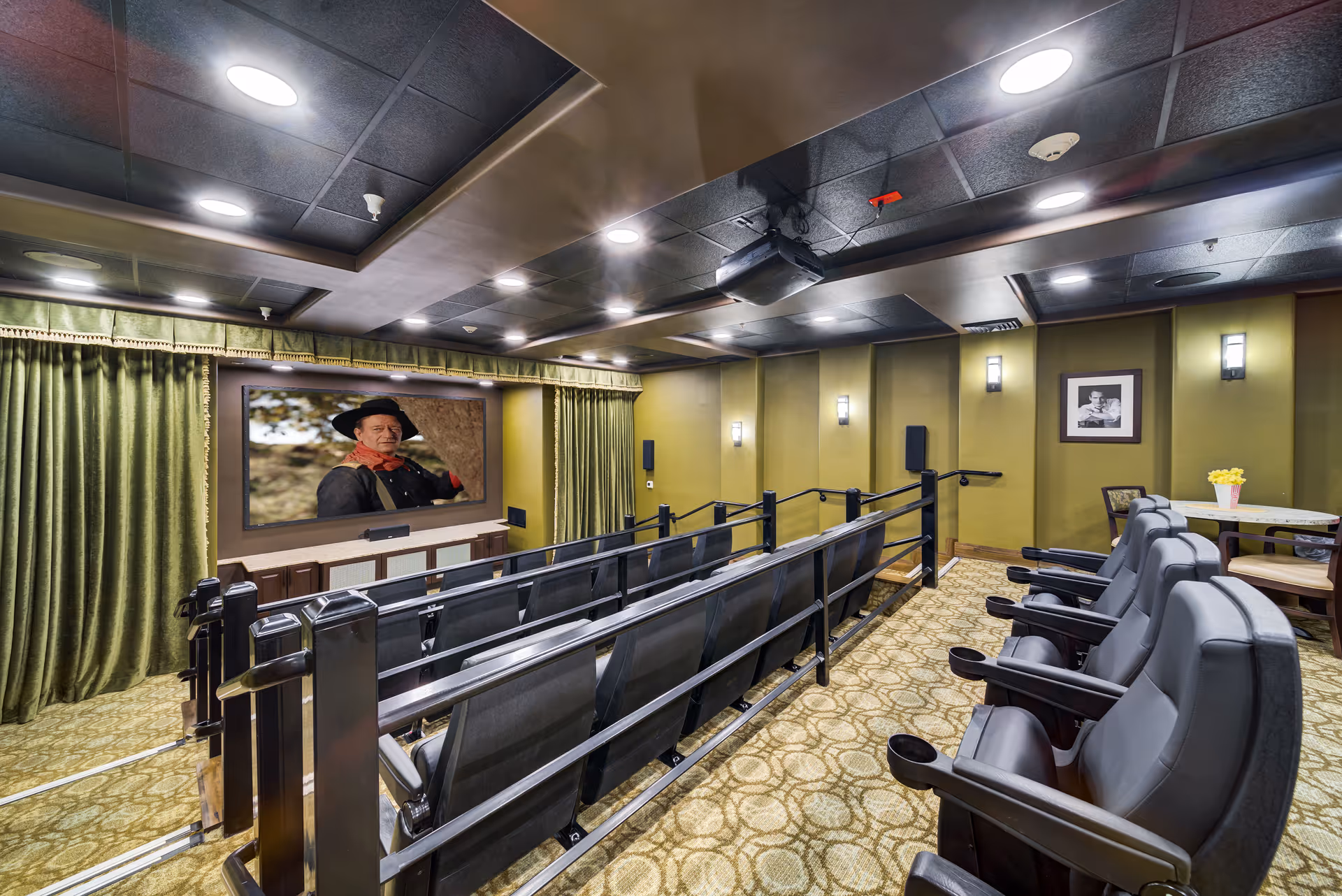 Interior view of a small theater room with two rows of black cushioned seats facing a large screen showing a man in a cowboy hat. The room has green walls, green curtains around the screen, patterned carpet, and a ceiling with recessed lighting. There is a small table with chairs and a vase of yellow flowers in the back corner.