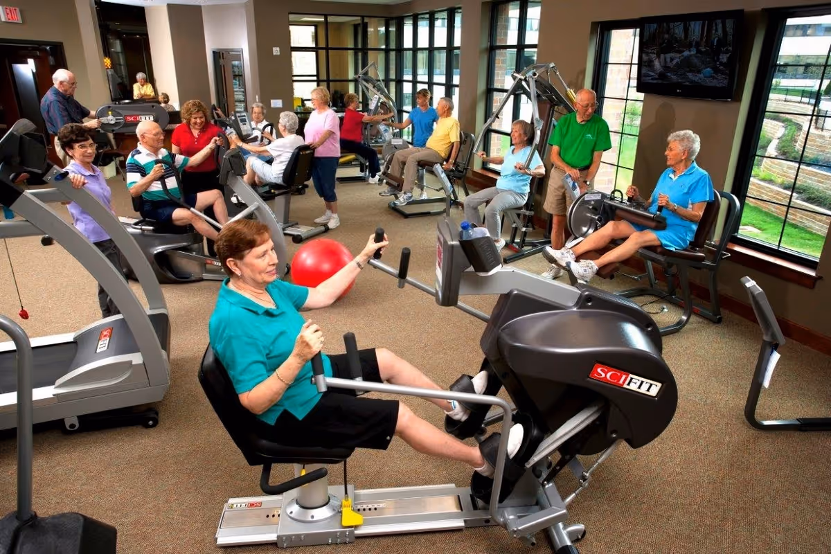 A group of elderly people exercising in a fitness room with various exercise machines. The room has large windows showing an outdoor garden area. The participants are using equipment such as rowing machines and stationary bikes, and appear engaged and active.