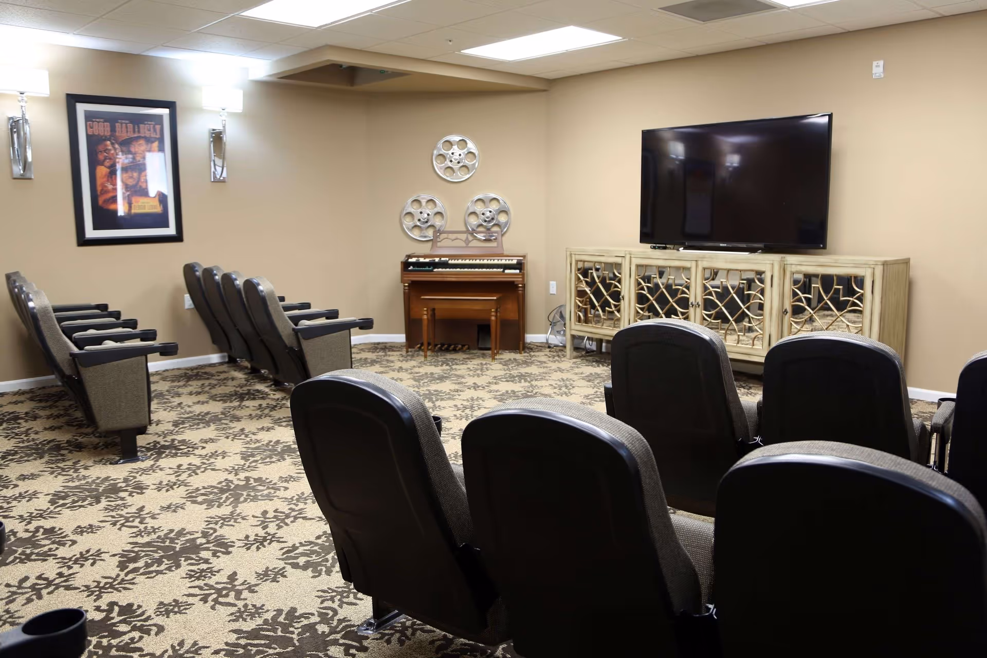 A small theater-style room with two rows of cushioned chairs facing a large flat-screen TV mounted on a decorative cabinet. The room has beige walls, patterned carpet, a framed movie poster on one wall, and a small organ with decorative film reels above it.