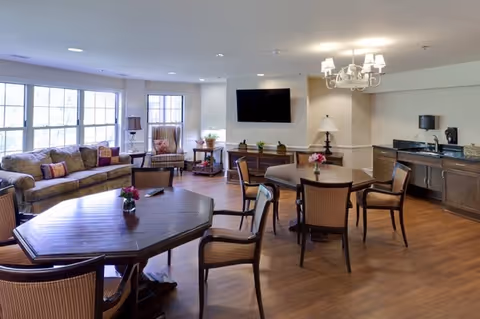 A spacious and well-lit common area with wooden floors, featuring several round wooden tables with chairs, a comfortable sofa with cushions, an armchair, a flat-screen TV mounted on the wall, and a small kitchenette area with a sink and coffee maker. The room has large windows allowing natural light to enter.